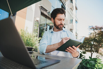 Young businessman writing in a notebook outdoors with a laptop at a trendy urban location