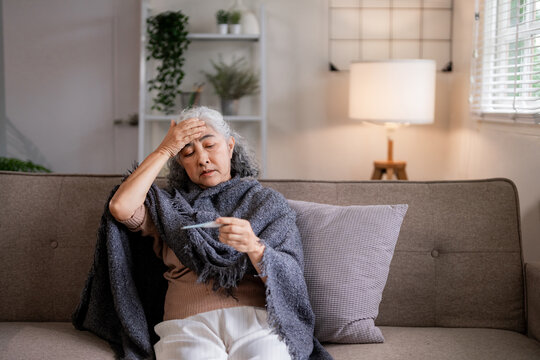 Asian senior woman unwell at home measures body temperature on couch blanket wrapped feeling fatigue and headache health concern for elderly wellness