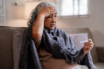Asian older woman ill fever checking temperature home senior health concern sofa domestic illness self care medical symptom discomfort indoor unwell elder health at home