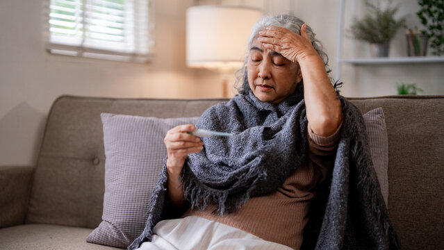 Older Asian woman sick home measuring temperature with thermometer on sofa ill feeling unwell fever health checkup disease symptoms self care recovery wellbeing discomfort