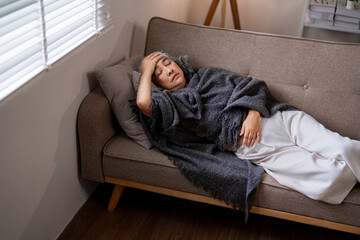 Older Asian woman resting unwell on couch at home experiencing headache pain fatigue discomfort...