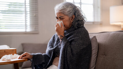 Older Asian woman blowing nose on sofa at home feeling unwell sick senior lady resting indoors...