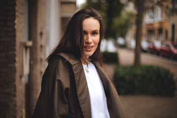 Fototapeta premium Woman wear trench coat. Portrait of young happy long hair brunette woman on city street looking at camera, laughing. Confident stylish girl outdoor. Proud and satisfied woman standing on street.