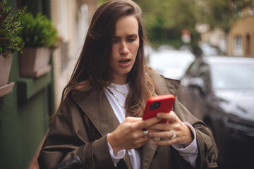 Outdoor shot of serious woman uses modern gadgets walks at street uses mobile phone, wears trench...
