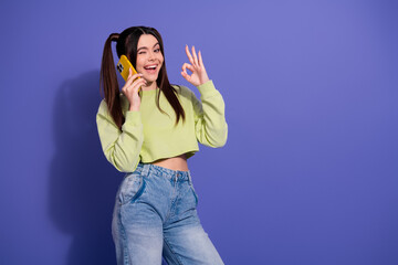 Smiling young woman with ponytails holding her phone while gesturing okay, casual trendy outfit on...