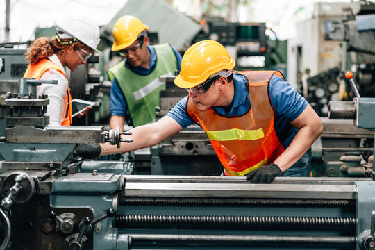Three factory workers are actively in large industrial machines. In the foreground, a male worker with a yellow hard hat and a bright orange safety vest is leaning over a lathe