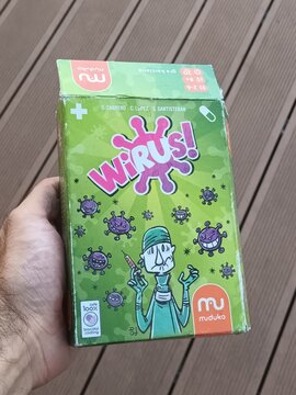 Hand holding a colorful green card game box titled 'Wirus!' with cartoon viruses and a doctor illustration on a wooden deck background