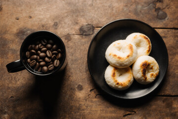 Rustic wooden table with black mug of dark coffee beans and black plate of golden-brown, fluffy donuts.