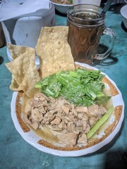 A close-up shot of a delicious bowl of Indonesian chicken noodles (Mie Ayam) topped with chopped chicken, green vegetables, and crispy fried wonton skins, served with a glass of iced tea.