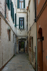 A narrow alleyway winds between old, weathered buildings, their rustic facades showing a history of wear. Old walls and green shutters frame the path. Venice, Italy keeps its secret charm.