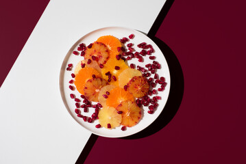 Rodajas de cítricos variados: naranja, limón, mandarina, naranja sanguina, lima y semillas de granada en un plato blanco sobre fondo granate. Vista superior