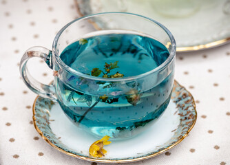 Glass cup filled with blue hot herbal tea, infused with dried flowers and herbs, placed on a decorative saucer. A warm, cozy beverage often enjoyed for its soothing and healthful properties