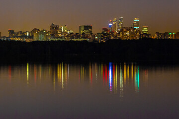 Fototapeta premium Night view of Strogino Backwater on Moscow River with reflections of city lights and Moscow-City skyscrapers silhouette against dark forest. Urban atmosphere. Russia.