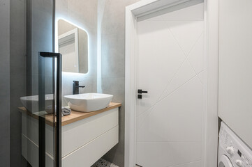 A clean, modern bathroom with a white vanity, vessel sink, illuminated mirror, and a white door with geometric designs. A washing machine is also visible on the right