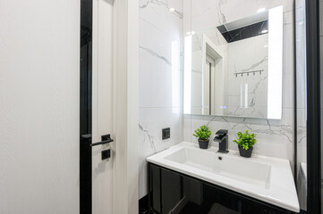 A modern bathroom with white marble tiles, black accents, a lighted mirror, and a stylish sink