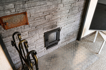 A textured gray stone wall features an ornate metal door, decorative plate, and fireplace tools....