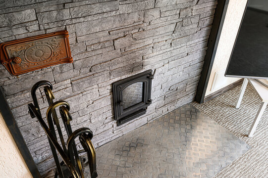 A textured gray stone wall features an ornate metal door, decorative plate, and fireplace tools. The floor is metal, leading to a carpet and furniture leg