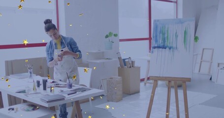 Holding smartphone, female artist in paint apron examining canvas drips at art studio, with easel