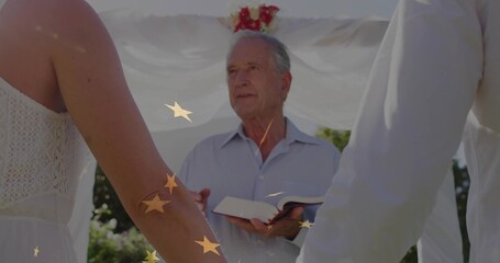 Conducting officiant reading ceremonial book under white draped arch in garden, with joined hands