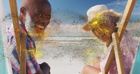 Smiling couple turning heads toward viewer on beach, with wooden chairs and straw sun hat
