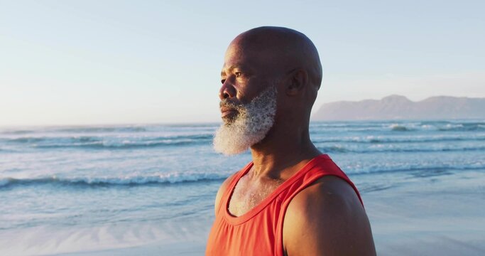 Standing senior man with white beard facing ocean at beach, with red sleeveless shirt, copy space