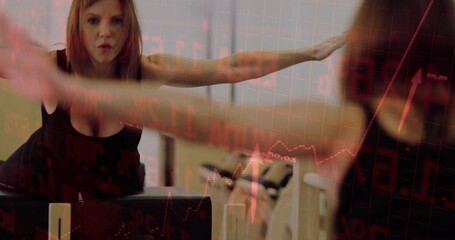 Extending arms woman wearing sports bra stretching in studio with mirror, reformer and red charts