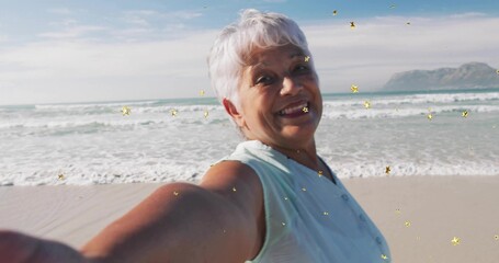 Smiling senior woman extending arm on sandy shoreline, with smartphone and star-shaped sparkles