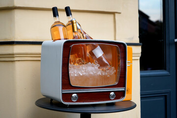 Vintage TV repurposed as an ice bucket holds chilled rose bottles on a small round table by a doorway. Playful upcycling scene mixing mid-century design with modern outdoor bar decor.