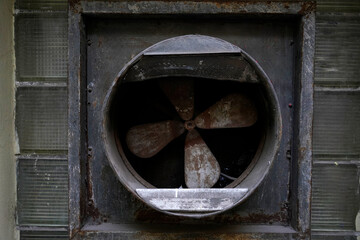 Rusty industrial exhaust fan set in a glass-block wall, weathered blades and soot-stained housing. Gritty urban detail showing ventilation, decay, and factory patina.