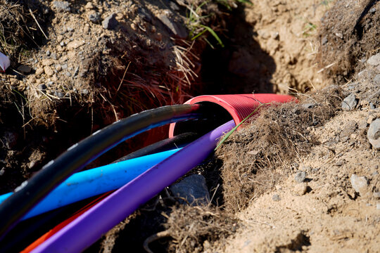 Black, blue, and purple utility cables feed into a red corrugated conduit within a narrow trench. Fresh soil and rocks frame a close view of underground installation for telecom and power networks.