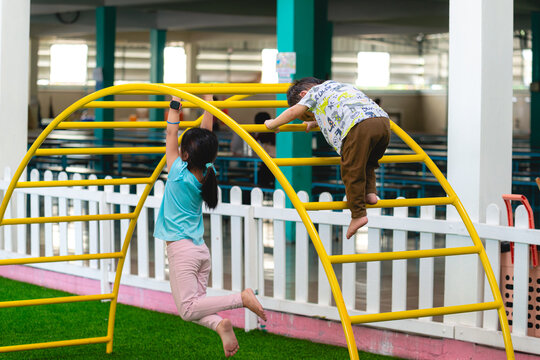 Candid of two young children actively playing on curved yellow climbing frame. Boy is scrambling over top while girl hangs from bar, fun and physical activity. Moment of active play and childhood joy.