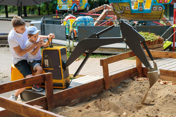 A senior boy helps a junior boy operate a yellow excavator at a children's amusement park on a...