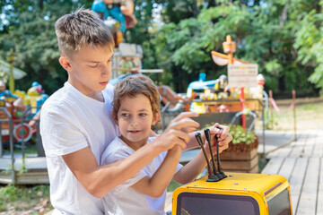 A senior boy helps a junior boy operate a yellow excavator at a children's amusement park on a...