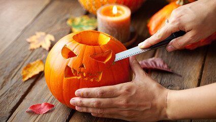 Close-up of hands carving a spooky face into a pumpkin for Halloween, with autumn leaves and a lit candle in the background. Festive seasonal preparation and holiday atmosphere.