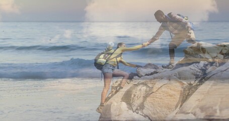Hikers wearing yellow shirt and plaid shirt climbing rocky outcrop by ocean waves, with backpacks