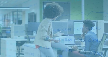 Collaborating women discussing project at open-plan office desk, with tablet and monitors