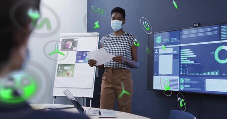 Businesswoman wearing face mask and blouse reading papers in meeting room, with flipchart and icons
