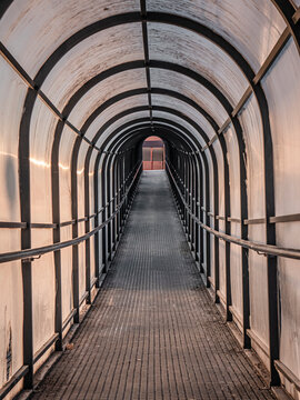 Pedestrian tunnel on an upward slope composed of a longitudinal arched metal structure of the same material and covered with a layer of translucent plastic through which the evening light enters
