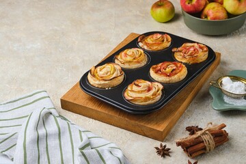Puff pastry with rose-shaped apples in a black muffin tin on a wooden board against a light concrete background.
