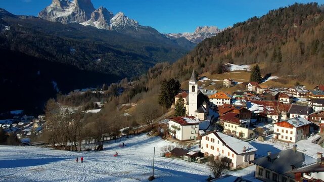 Winter Family Activities in Vinigo di Cadore near Cortina d'Ampezzo: Children Sledding on Snowy Hill by Historic Alpine Church, Aerial 4K Drone View of Dolomites Italy 2026 Winter Olympic Venue Region