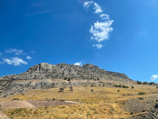 Rocky mountain slope under a bright blue sky with scattered clouds in Greece. Dry yellow grass and rugged stone landscape on a sunny summer day. Natural Mediterranean scenery and geology view.