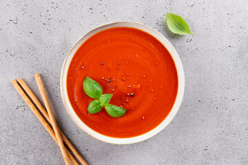 Tomato cream soup with fresh basil and breadsticks on a light stone background. Top view. Healthy vegetarian meal.