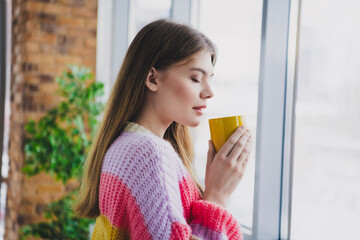 Woman enjoys a warm drink by the window wearing a cozy pastel sweater
