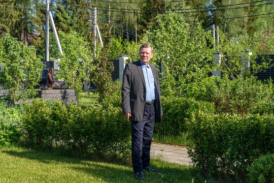 Composed middle-aged man in dark suit jacket and blue shirt stands attentively in sun-drenched, lush green garden filled with abundant foliage and distant trees.