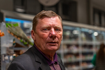 Intense close-up portrait of older Caucasian man with blue eyes, wearing dark suit jacket and purple polo, gazing directly at viewer amidst blurred supermarket aisle background.
