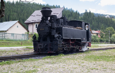 vintage steam locomotive from the Mocănița narrow-gauge railway in Romania, gently moving along the tracks through a scenic rural landscape with forested hills and houses in the background
