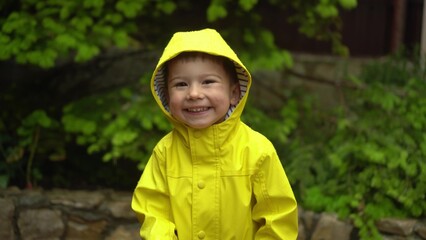 child in a yellow raincoat with hood smiles in the rain with green garden background