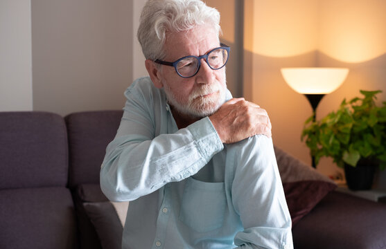 Senior man sitting on sofa at home holding his shoulder with a painful expression, suffering from joint pain or muscle discomfort. Concept of aging, arthritis, injury, or health problems