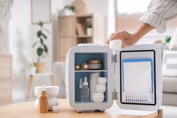 Young woman with jar of cosmetic product and small refrigerator on table at home