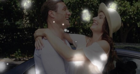 Hugging couple laughing in driveway under sunlight, wearing dress, straw sun hat beside parked car